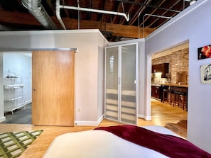 Un interno moderno con un letto, una porta in legno, un angolo cottura con parete in mattoni e un'area pranzo con sgabelli da bar.