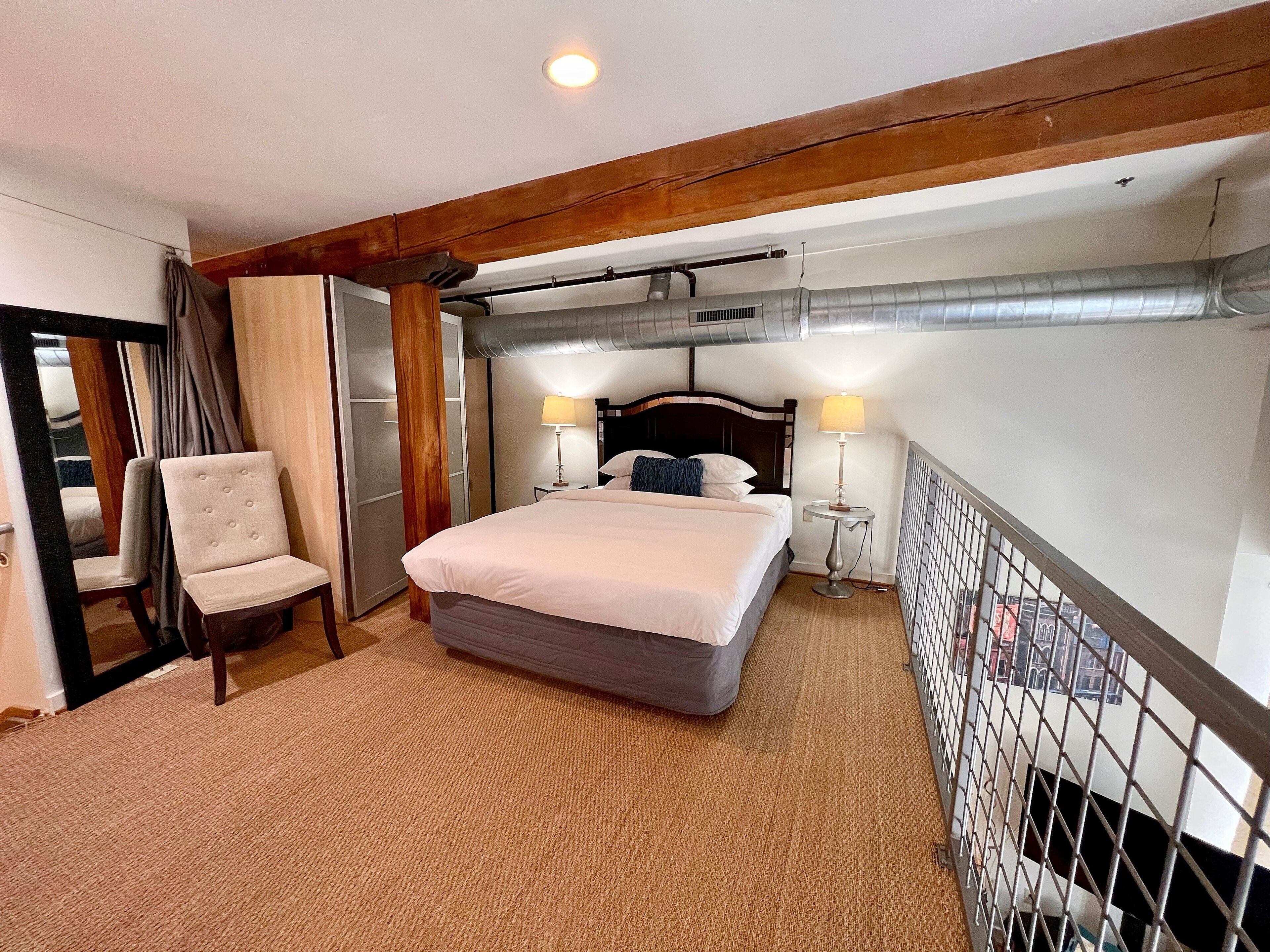 A hotel room with a bed, two bedside lamps, a chair, a mirror, and a view of the outside.
