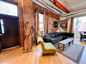 A modern living room with a brick wall, a black sofa, a yellow armchair, a wooden sculpture, and a dining area with a table and chairs. - Dunn (Nashville)