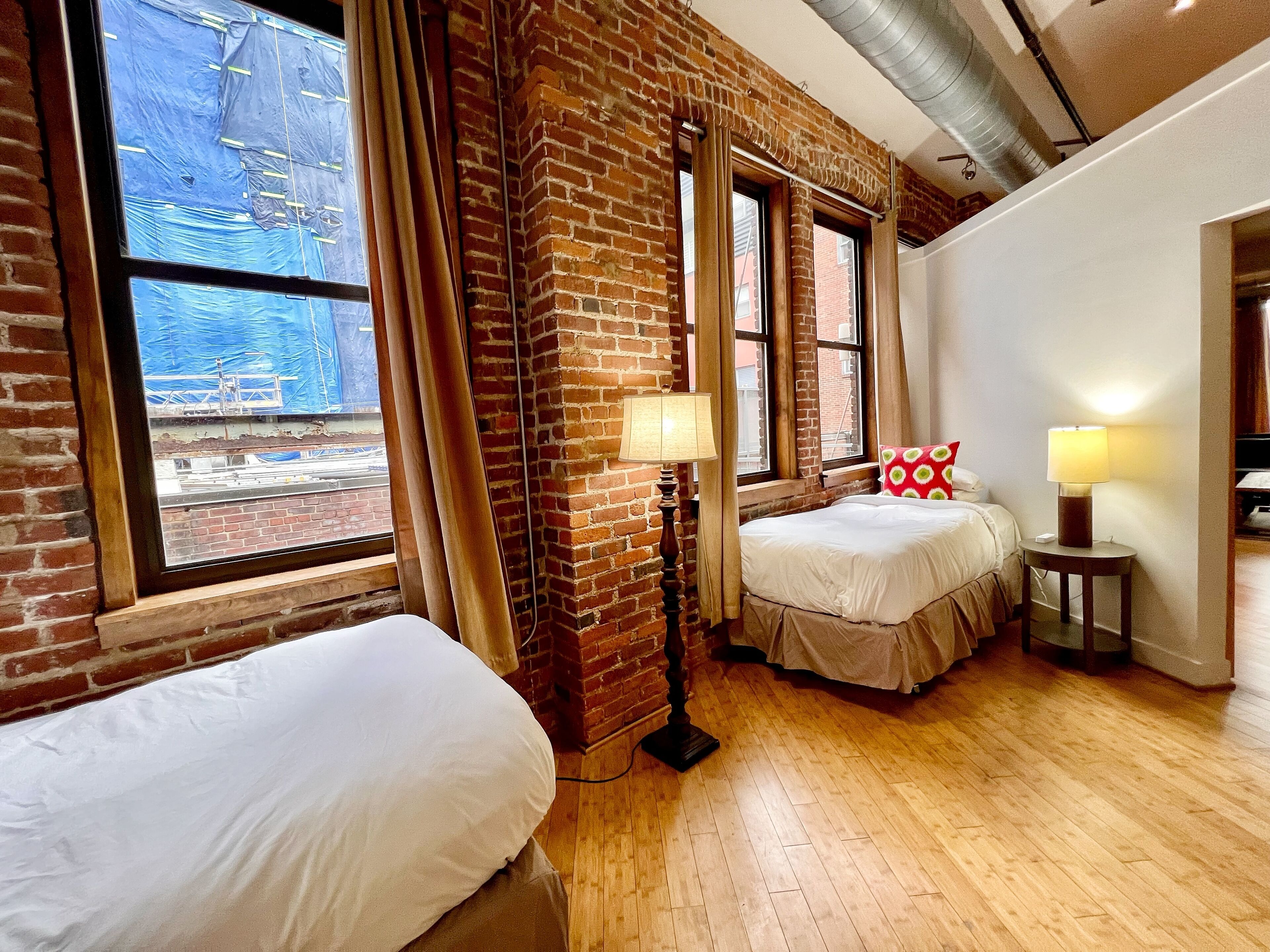 A room with two beds, a brick wall, large windows, and a wooden floor.