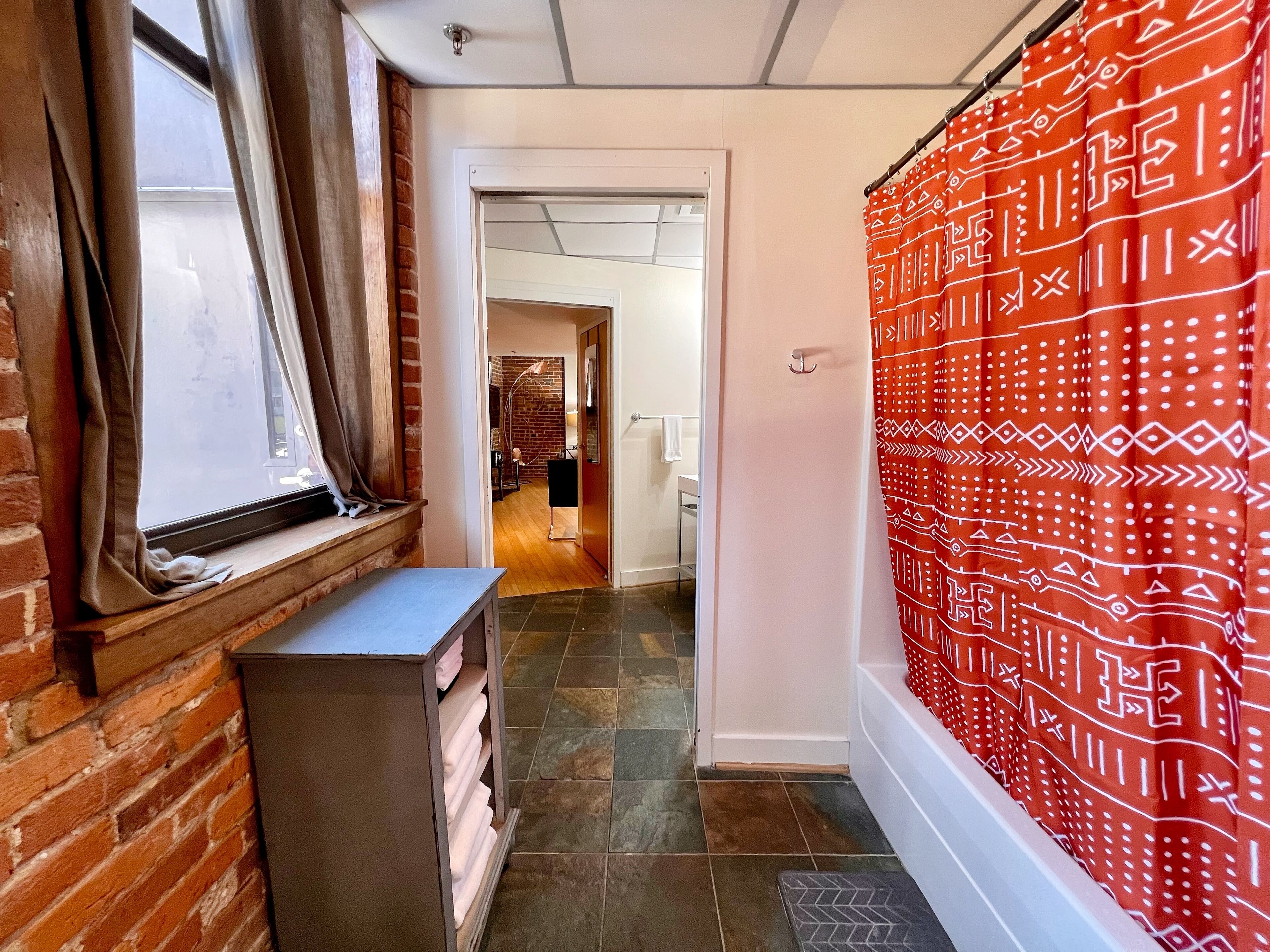 Een gang met een bakstenen muur, een raam met gordijnen, een rood gordijn met patroon, en een betegelde vloer.