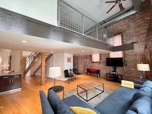 A spacious living area with a high ceiling, a staircase leading to an upper level, a large sofa, a coffee table, and a flat-screen TV mounted on the brick wall. - Elvis (Nashville)