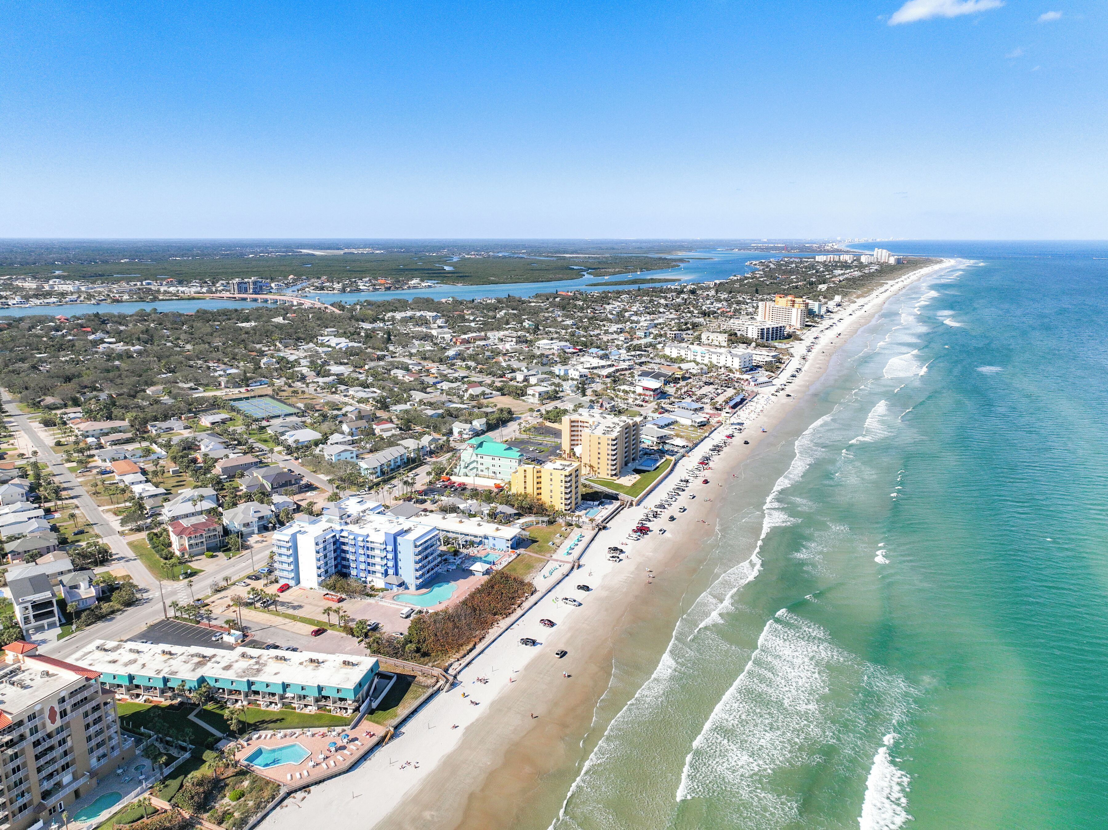 An aerial view of a coastal city with a long beachfront, dense residential area, and several hotels with multiple floors and balconies.
