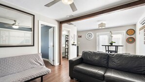 A living room with a black leather sofa, a grey quilted sofa, wooden flooring, a ceiling fan, and a dining area with a table and chairs. - Central Phoenix Casita Unit C (Phoenix)