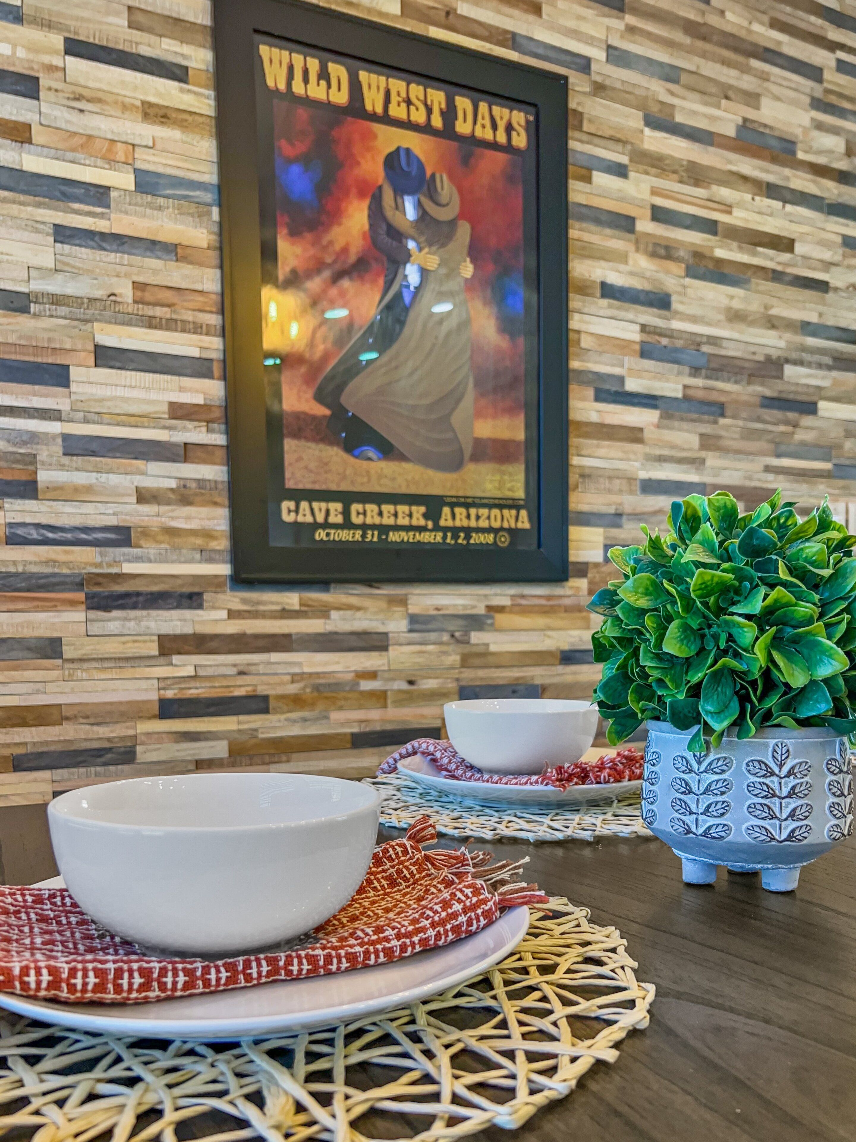 A framed poster of 'Wild West Days' in Cave Creek, Arizona, with a rustic wooden wall background and a table with a bowl and a potted plant.