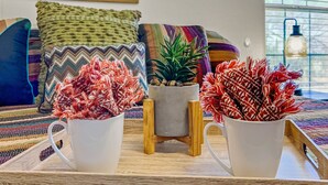 Ein Wohnzimmer mit einer Couch, bunten Kissen, einem gerahmten abstrakten Kunstwerk und Zimmerpflanzen in weißen Töpfen.