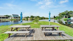 A wooden deck with picnic tables and umbrellas overlooking a calm lake, surrounded by greenery and buildings. - 8820 Rocky Creek Drive Unit A (Tampa)