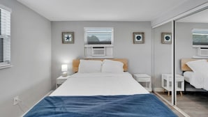 A modern bedroom with a bed, bedside tables, a mirror, and a window with blinds. - 8820 Rocky Creek Drive Unit A (Tampa)