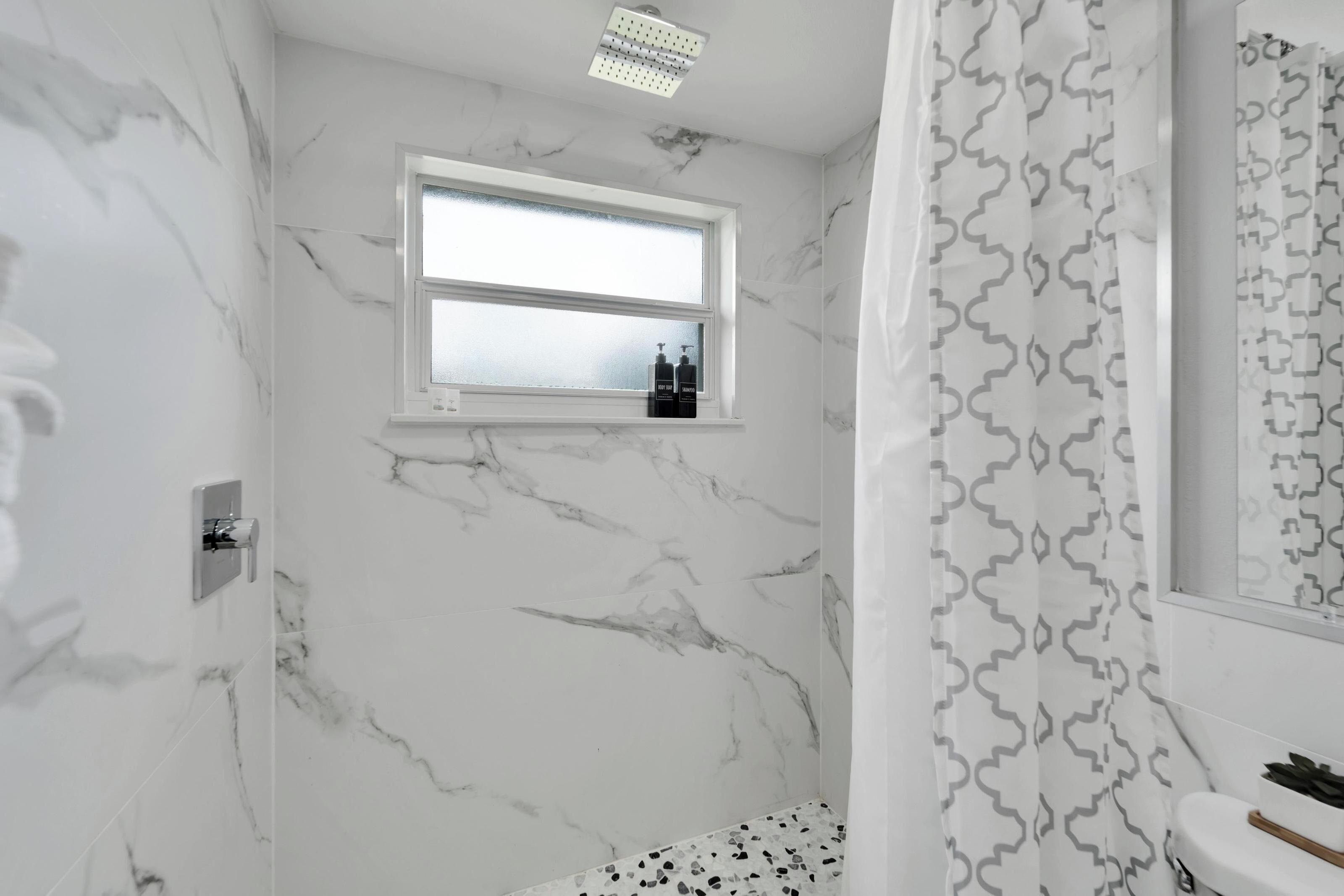 A bathroom with marble walls, a window with bottles, a shower curtain, and a toilet.