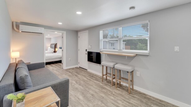 A compact living space with a sofa, a small coffee table, a kitchen area with a countertop and stools, and a window with blinds. - 8820 Rocky Creek Drive Unit B (Tampa)