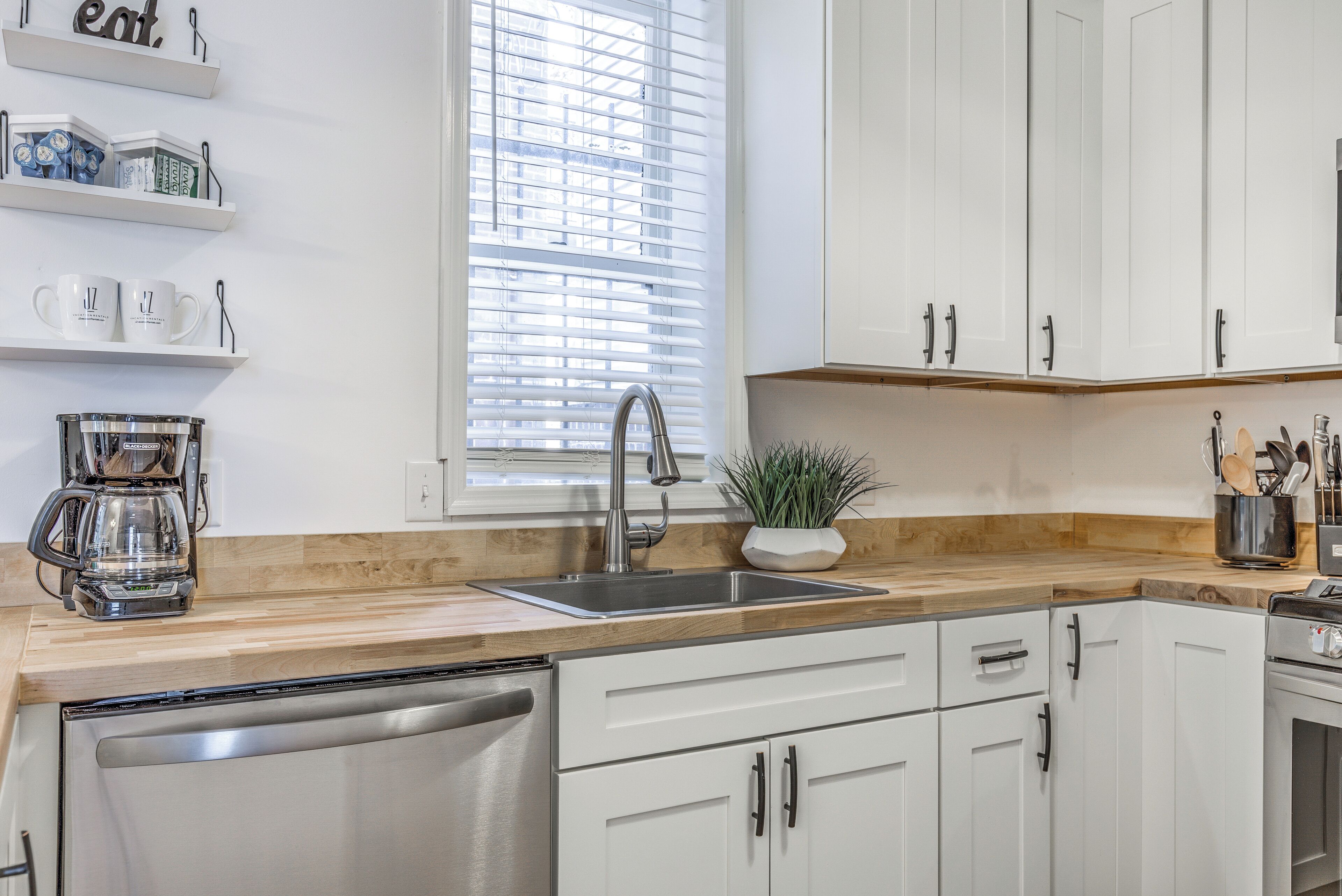 A modern kitchen with white cabinets, a stainless steel dishwasher, a coffee maker, and a window with blinds.