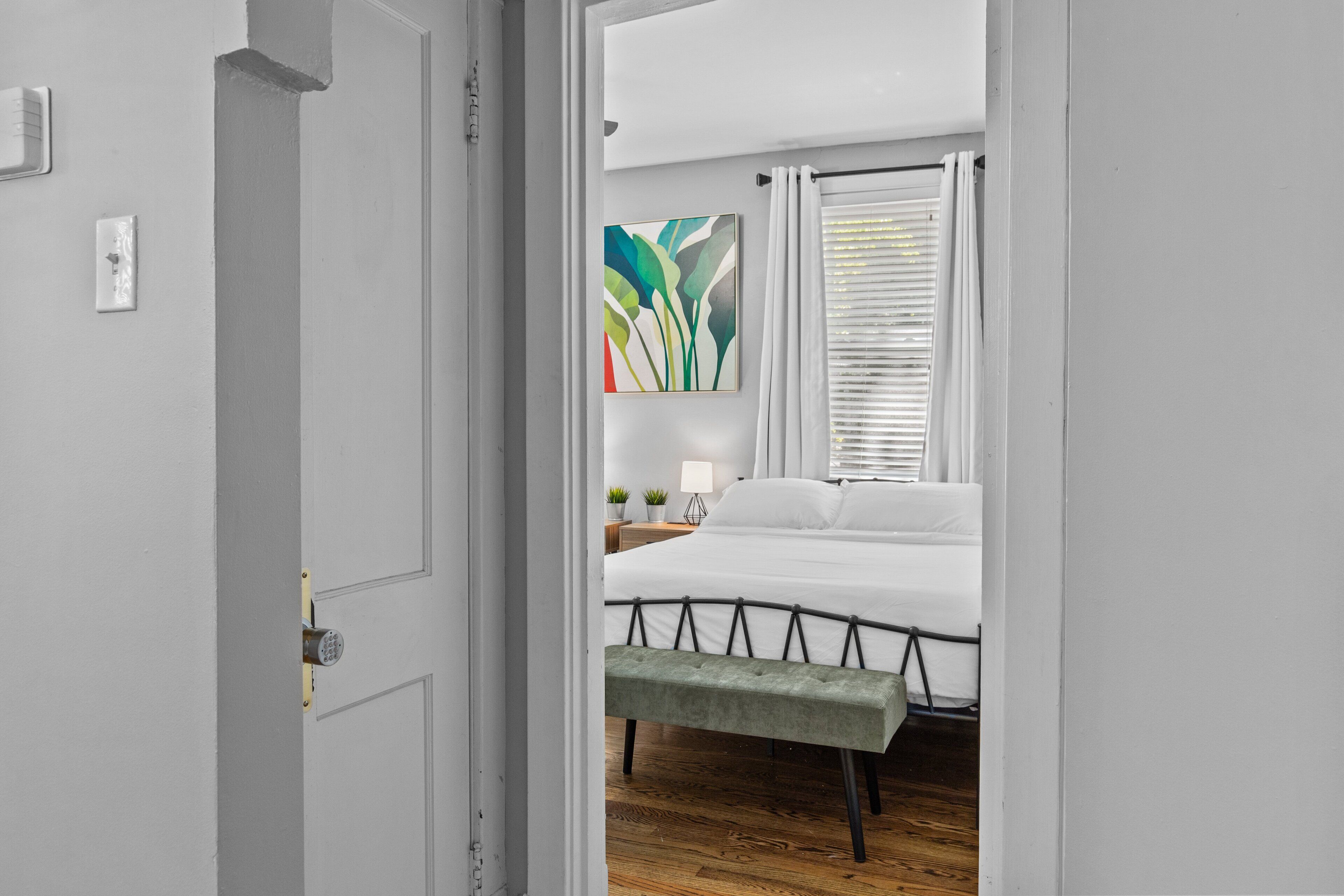 A bedroom with a bed, a bench, a nightstand, a lamp, and a wall-mounted plant.