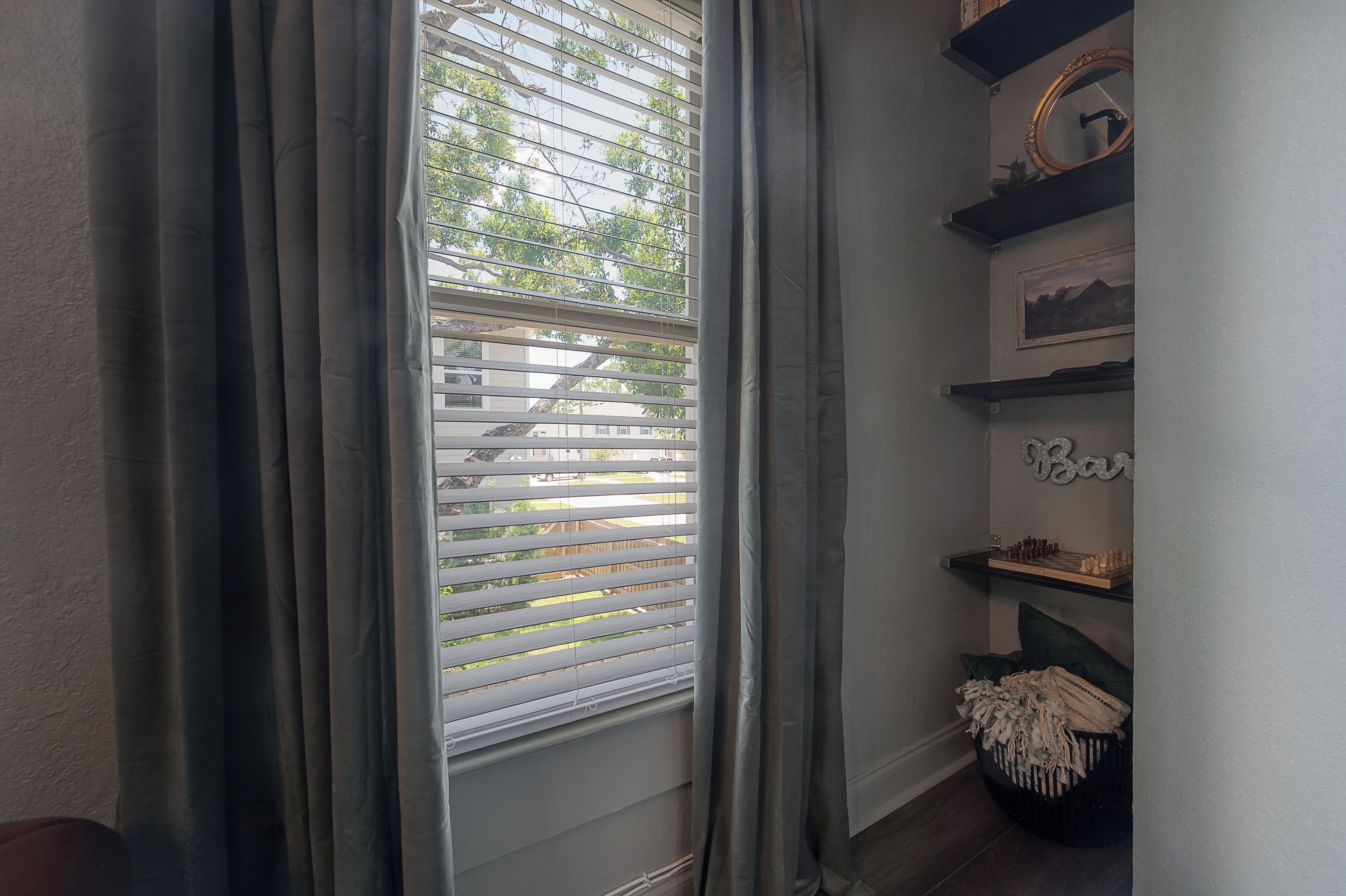 A room with a window with blinds, a shelf with decorative items, and a wall-mounted mirror.