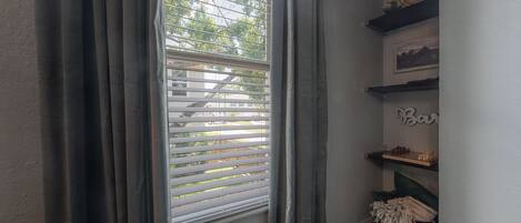 A room with a window with blinds, a shelf with decorative items, and a wall-mounted mirror.