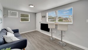 Een moderne woonkamer met een grijze bank, twee witte barkrukken en een groot raam met jaloezieën.