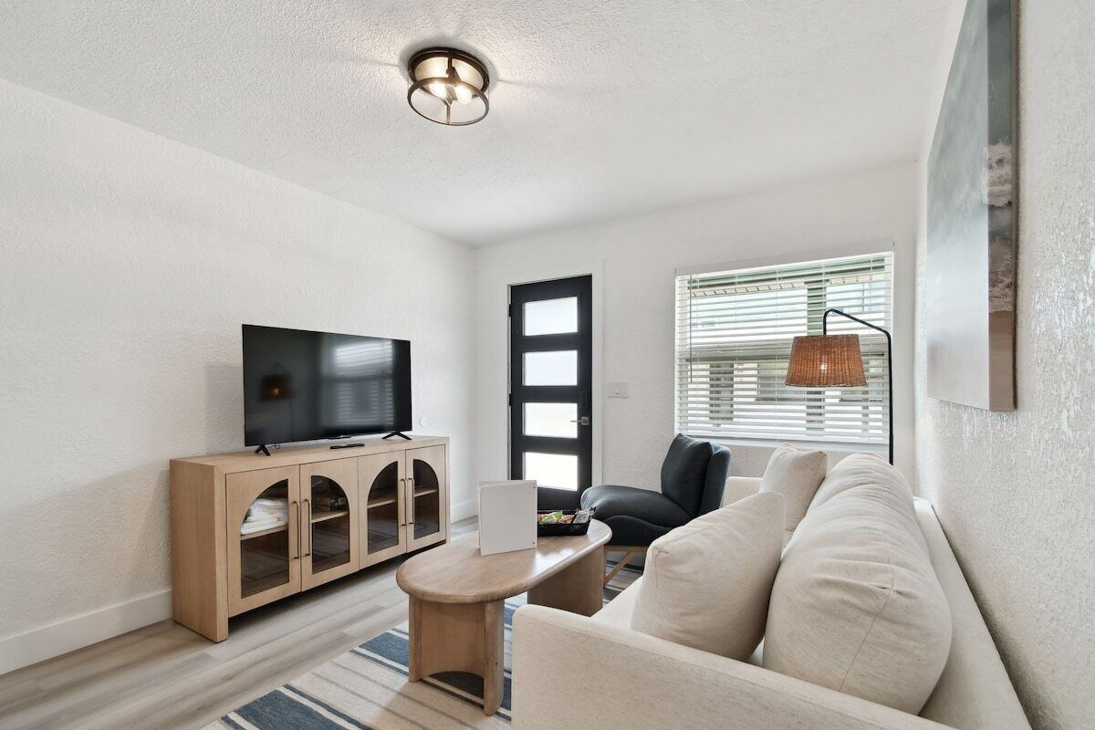 A modern living room with a flat-screen TV, a wooden cabinet, a sofa, and a window with blinds.