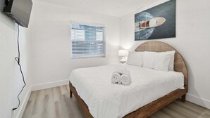 Una camera d'albergo con un letto, una TV fissata al muro, una finestra con persiane e un quadro raffigurante una barca.