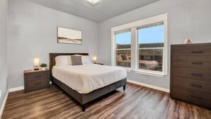 A bedroom with a large bed, a nightstand with a lamp, a dresser, and a window with a view of buildings. - Walk to DT Fast Wi-fi + Long Stay Equipped! (Tacoma)