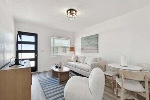 A modern living room with a sofa, coffee table, and dining area. There is a desk with a computer and a lamp. A painting is on the wall, and a window with blinds is visible. - Boardwalk John's Pass (Treasure Island)