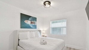A neatly made bed with white linens, a painting of a boat on the wall, and a window with blinds. - New Treasure Island Gem - Unit 3 (Treasure Island)