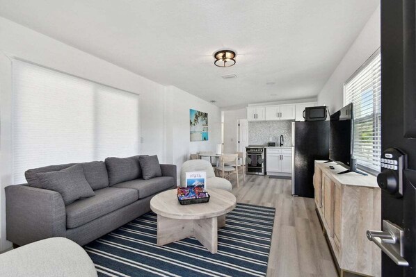 A modern living room with a grey sofa, a round coffee table, and a striped rug. There is a kitchen area with white cabinets and a black refrigerator. - Stylish Treasure Island Getaway - Unit 8 (Treasure Island)