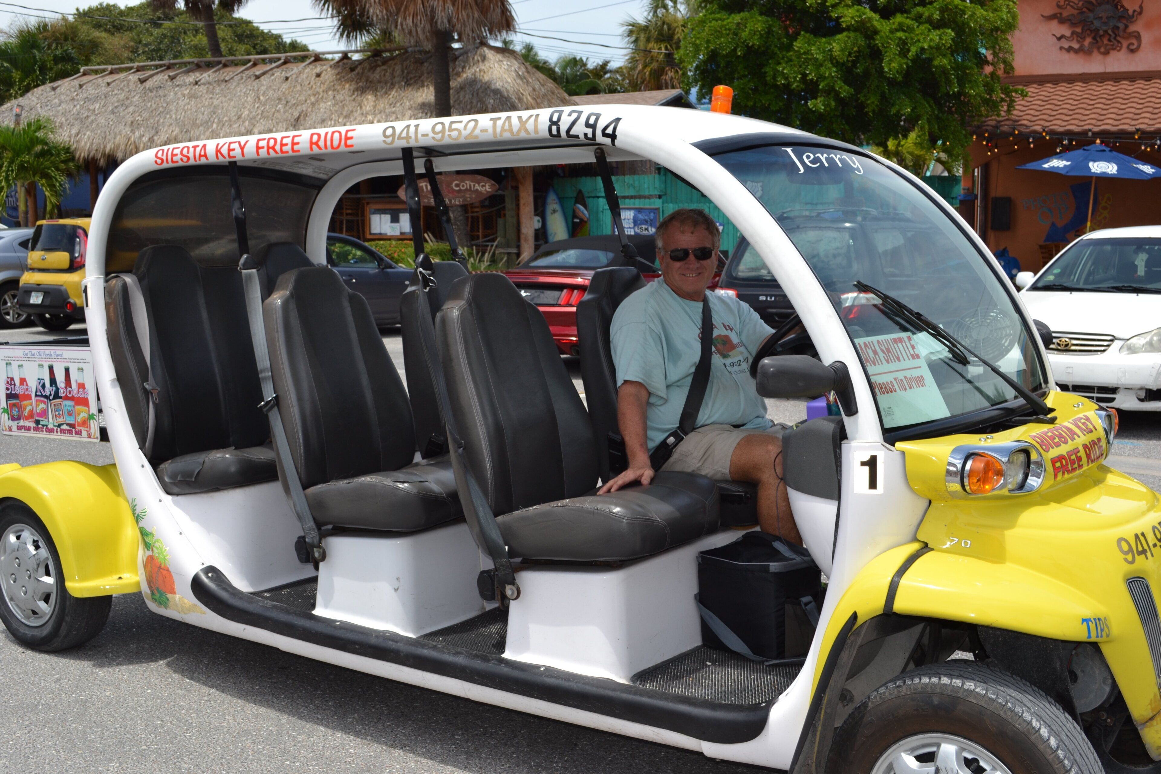 A compact, single-decker vehicle with a white and yellow color scheme, featuring a "Siesta Key Free Ride" sign and a contact number.