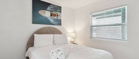 A neatly made bed with white linens and rolled towels, a headboard with a striped pattern, a painting of a boat on the wall, and a window with blinds.