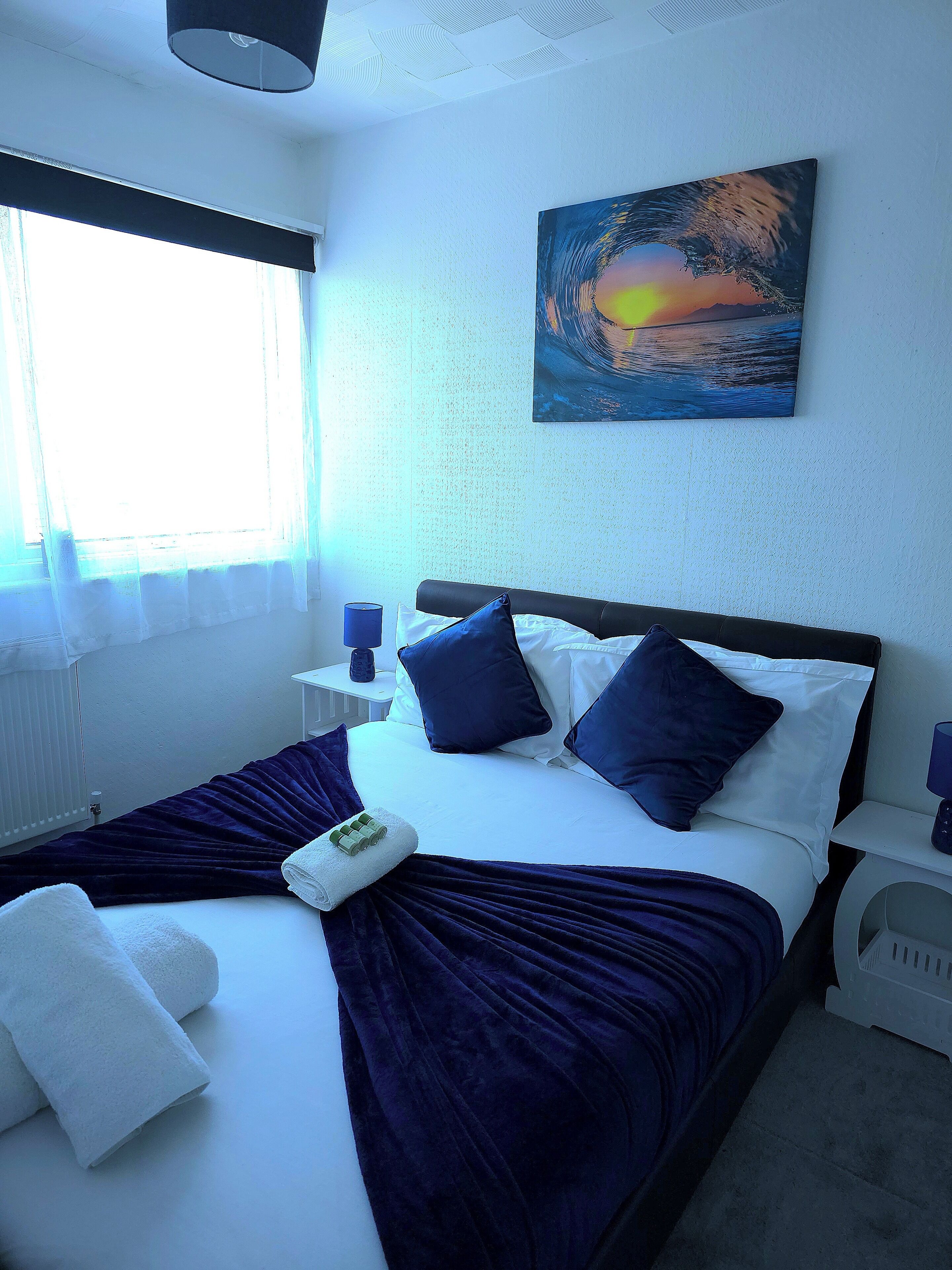 A hotel room with a bed, a painting of a sunset, a window with curtains, and a nightstand with a lamp.