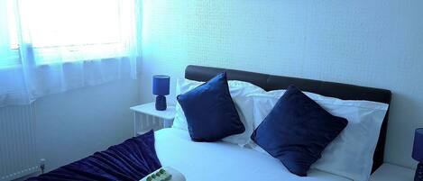 A hotel room with a bed, a painting of a sunset, a window with curtains, and a nightstand with a lamp.