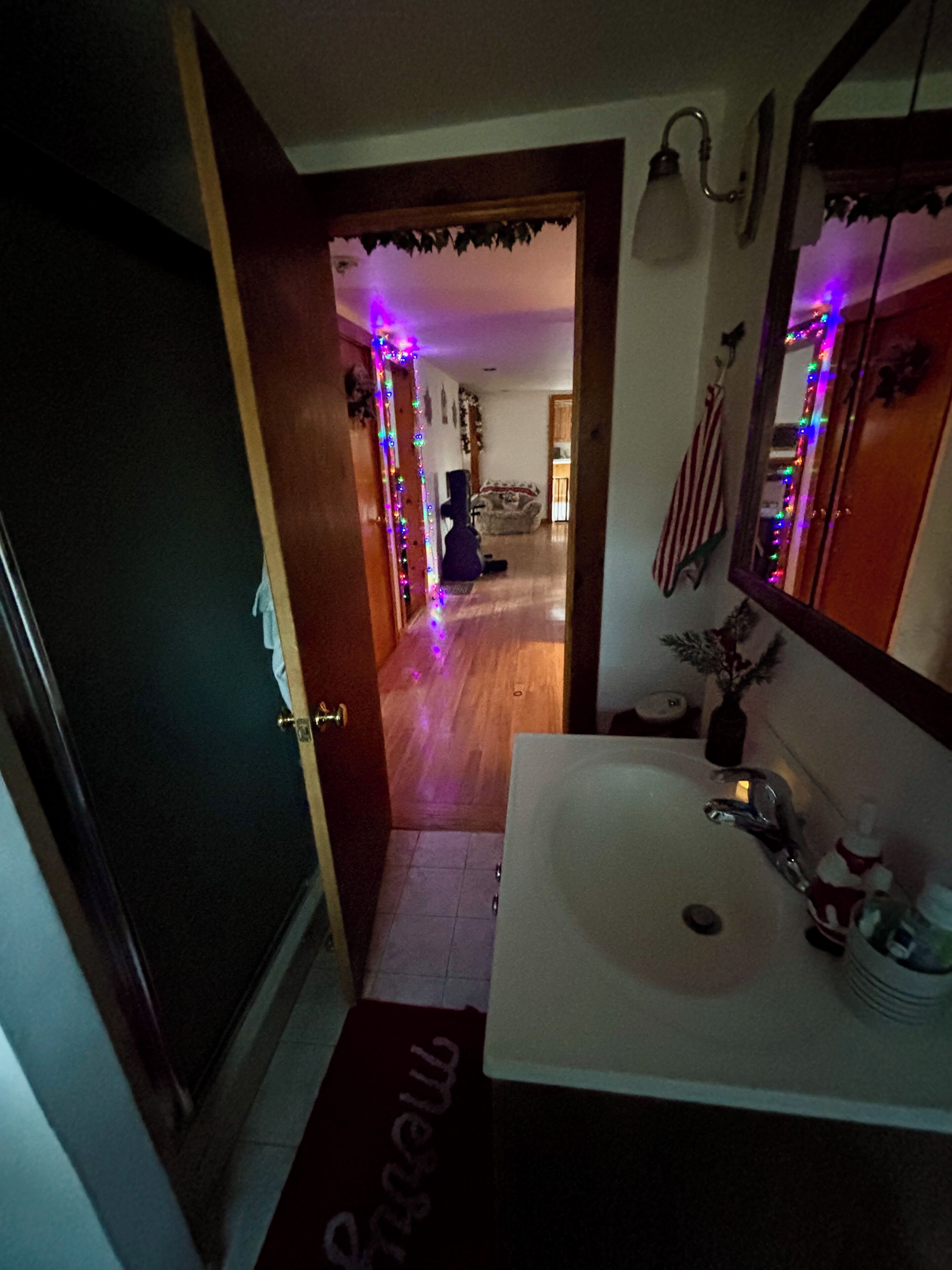 Un couloir étroit mène à une pièce où se trouvent un lavabo blanc et un miroir. Le couloir est décoré de guirlandes lumineuses colorées.