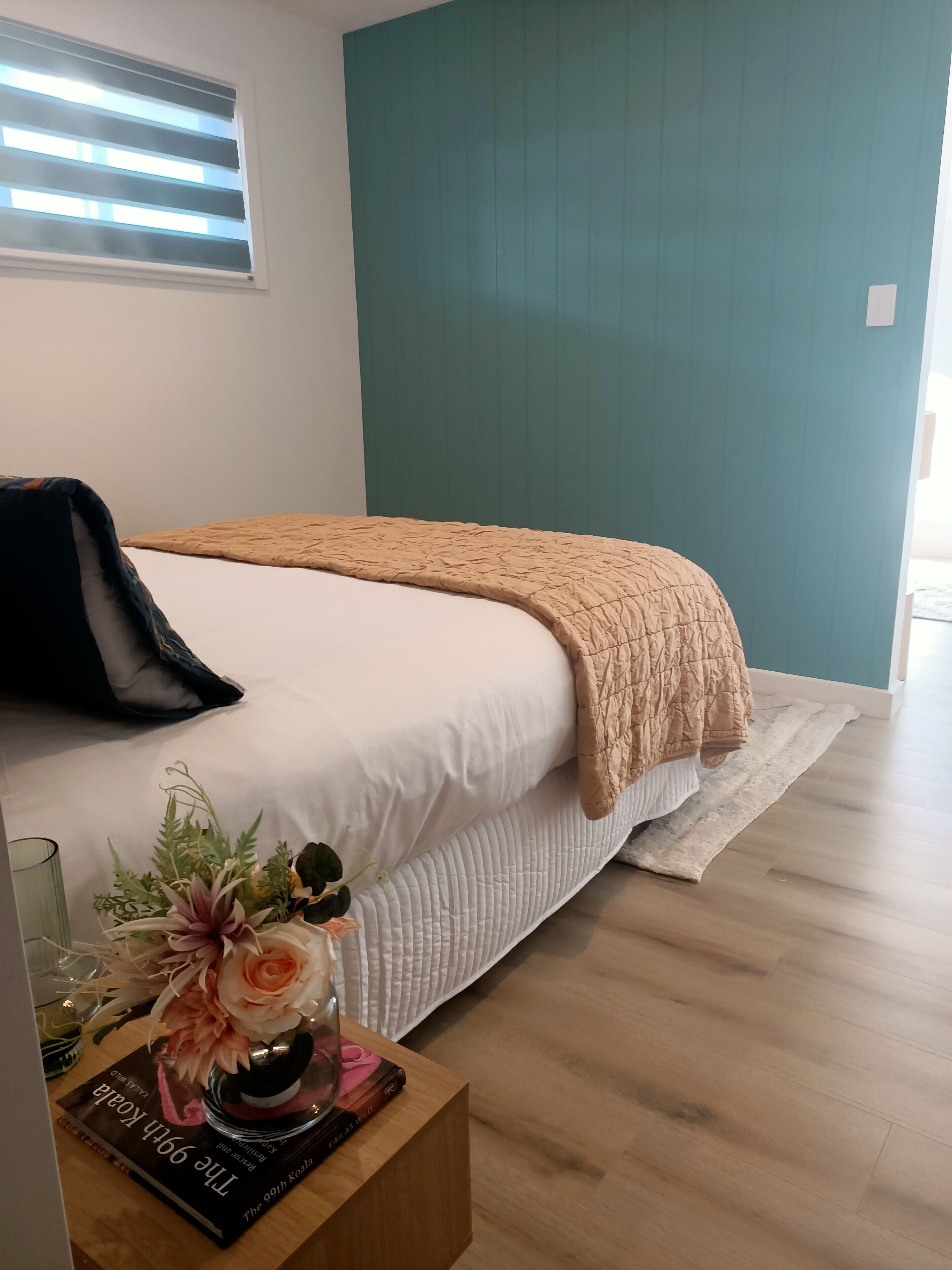 A neatly made bed with a beige blanket, a bedside table with a book and a vase of flowers, and a teal-colored wall.