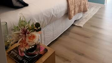 A neatly made bed with a beige blanket, a bedside table with a book and a vase of flowers, and a teal-colored wall.