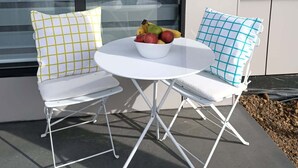 A white outdoor seating area with two chairs and a table, a bowl of fruit on the table, and a view of a building through large windows. - Cosy 1 Bedroom Unit with separate living space, laundry and kitchen. (Prospect Vale)