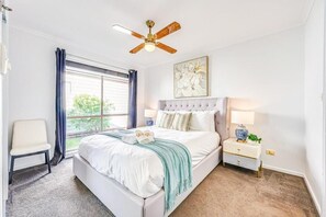 Un dormitorio con cama, ventilador de techo, ventana con cortinas, mesita de noche, una silla y un cuadro en la pared.