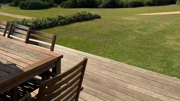Een houten terras met een tafel en stoelen, met uitzicht op een kustlandschap met een strand en begroeiing.