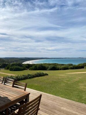 Une terrasse en bois avec une table et des chaises, offrant une vue sur un paysage côtier comprenant une plage et de la végétation.