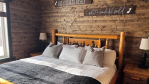 A wooden bed with a headboard, multiple pillows, and a blanket. There are two bedside tables with lamps. The wall has wooden panels and two signs with text.