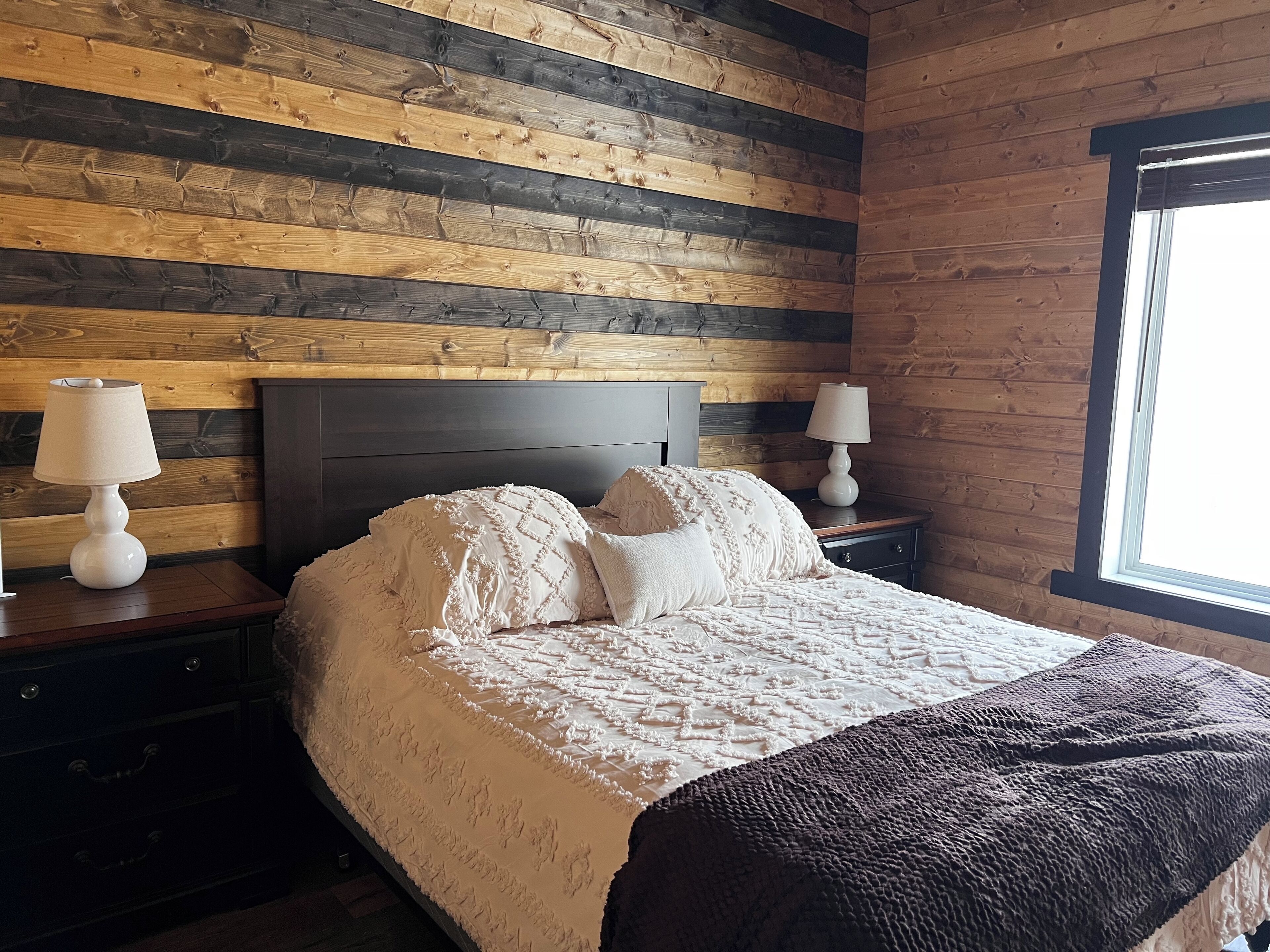 A bedroom with a wooden slat wall, a dark headboard, two bedside tables with lamps, a bed with a white bedspread and pillows, and a window on the right.