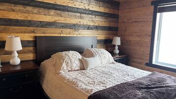 A bedroom with a wooden slat wall, a dark headboard, two bedside tables with lamps, a bed with a white bedspread and pillows, and a window on the right.