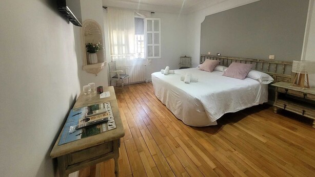 A bedroom with a wooden floor, a bed with white bedding, a bedside table, a chair, a window with curtains, and a small table with a book and a lamp. - Rural house in the heart of De la Vera in Yuste, Imperial retreat (Cuacos de Yuste)
