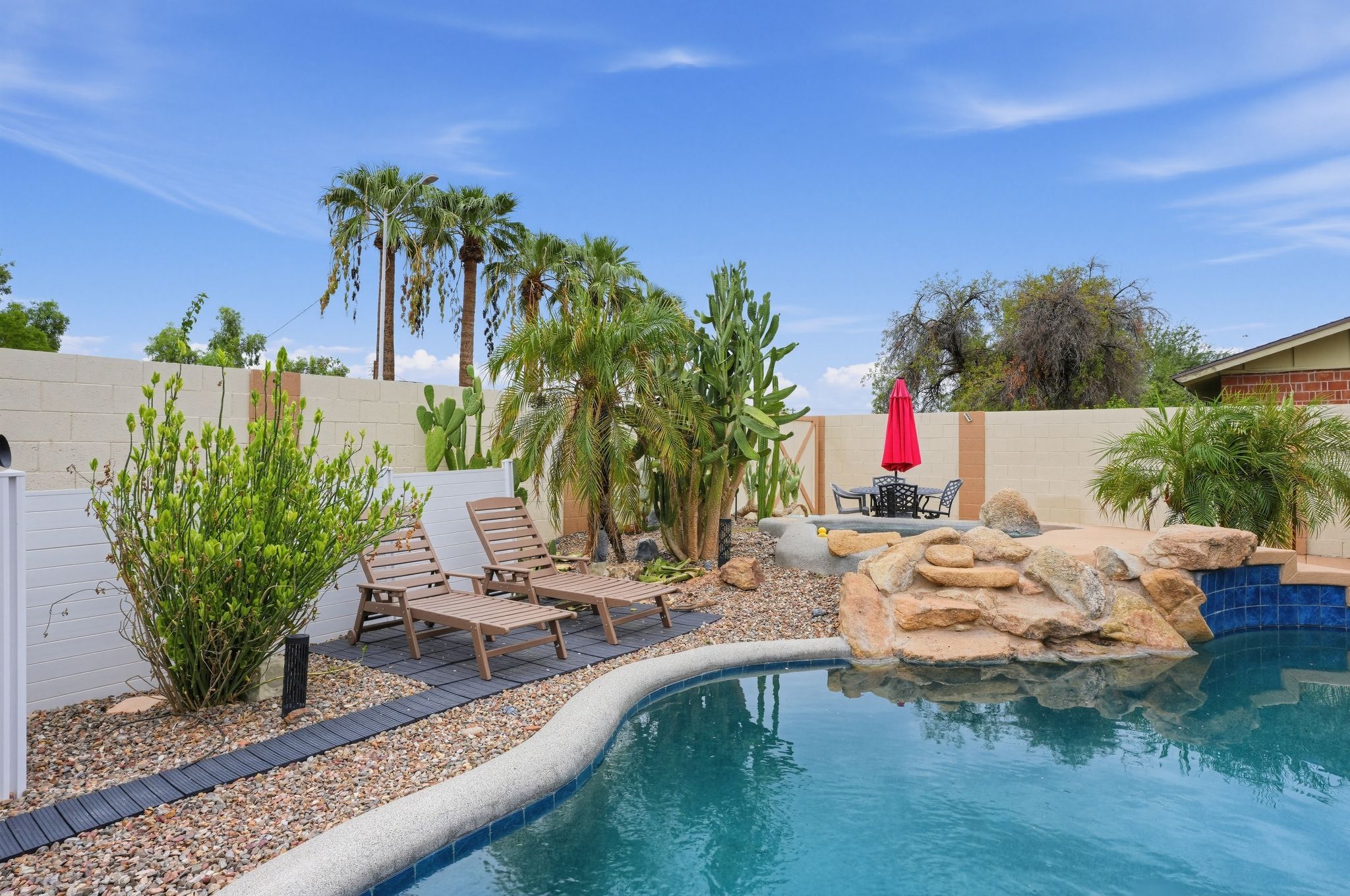 A swimming pool with lounge chairs and a red umbrella, surrounded by cacti and palm trees.