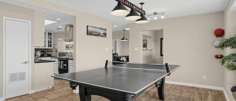 A ping pong table in a room with wooden flooring, a kitchen in the background, and a ceiling fan.