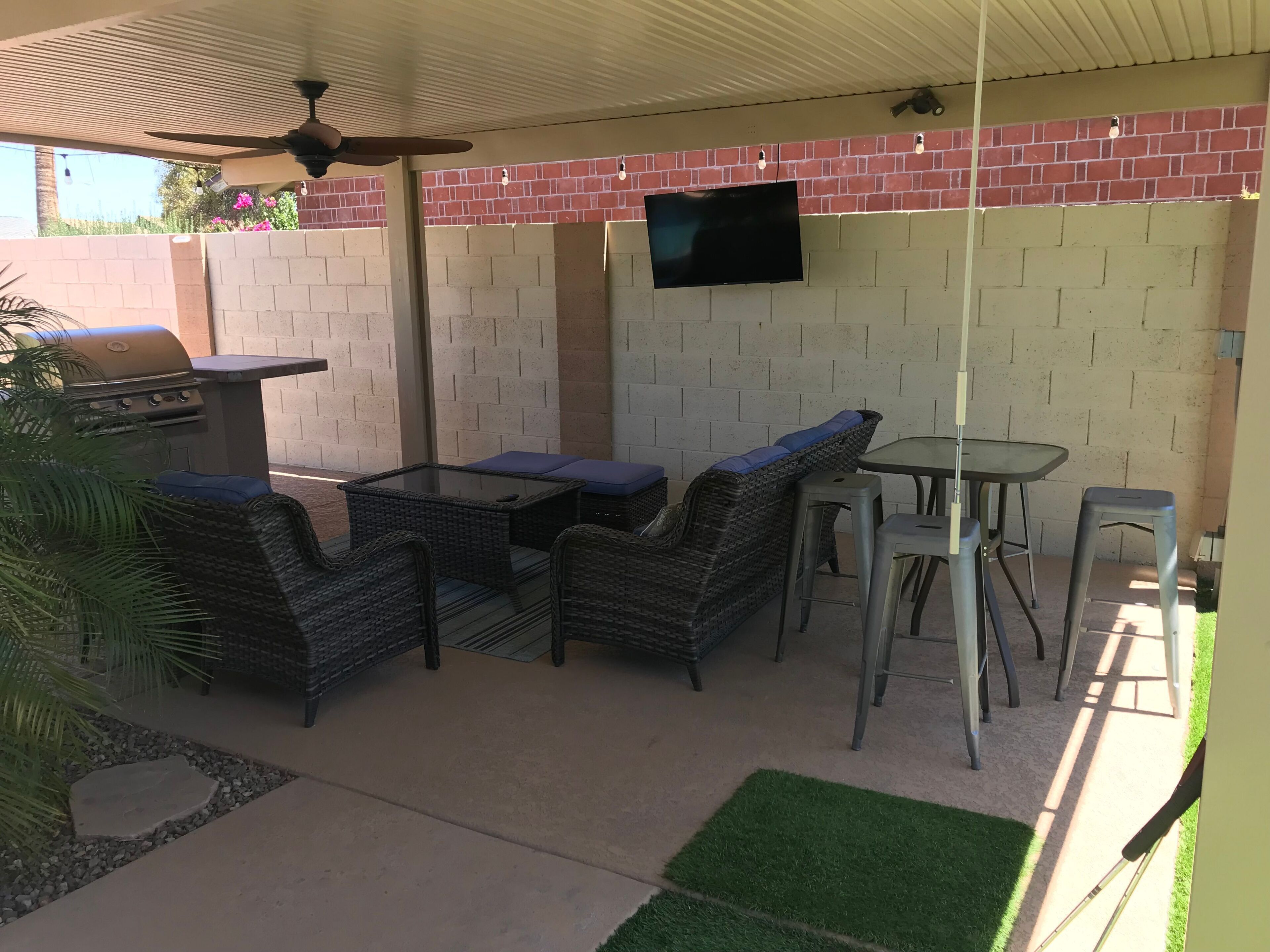 An outdoor patio area with a grill, wicker furniture, a table, and a TV mounted on a brick wall.