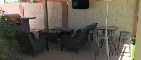 An outdoor patio area with a grill, wicker furniture, a table, and a TV mounted on a brick wall.