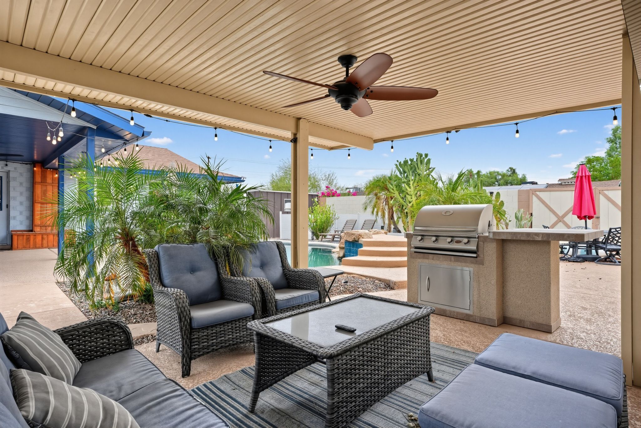Un patio exterior con mobiliario de mimbre, ventilador de techo, parrilla y piscina.