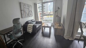 Uma sala de estar moderna com um sofá, uma mesa pequena e uma cadeira. Há uma janela com vista para edifícios.