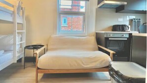 A compact room with a bunk bed, a small bedside table, a window with a view of a brick building, and a kitchenette with a stove and oven. - Golder Greens London City Bliss Apartment (London)