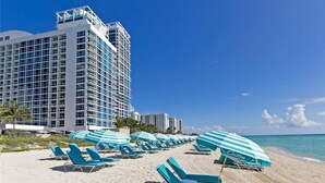 Une plage bordée de rangées de chaises longues et de parasols bleus, un grand bâtiment d’hôtel et un ciel bleu dégagé.