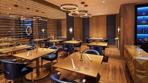 Un restaurante moderno con suelo de madera, sillas tapizadas en azul y una pared de ladrillo con una sección iluminada en azul. Las mesas están dispuestas con cubiertos y vasos.