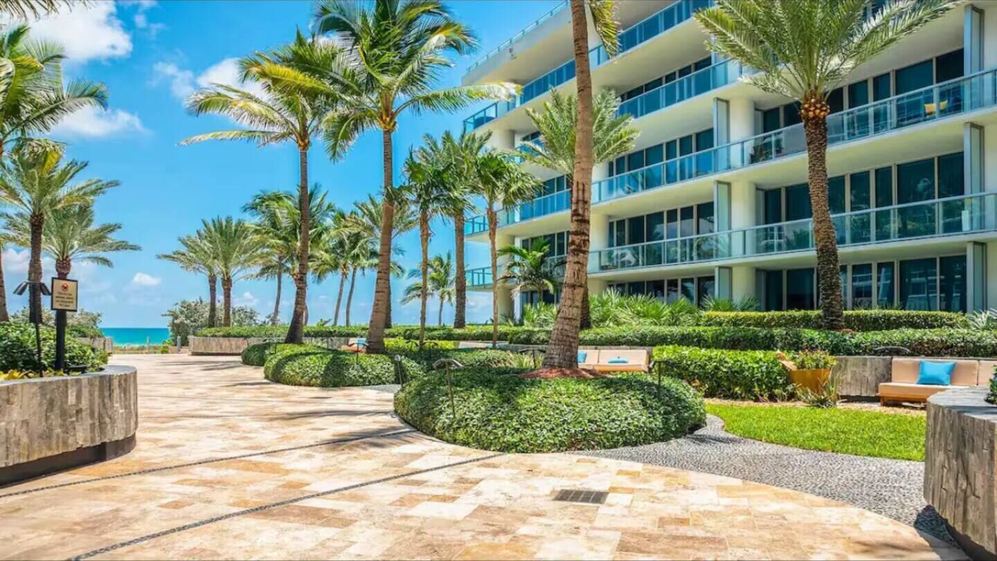 A resort with a palm-lined pathway leading to a modern building with balconies overlooking a beach.